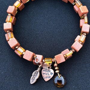 May include: A copper-toned bracelet with a heart charm that says "Made with Love". The bracelet is made with square beads and small glass beads.