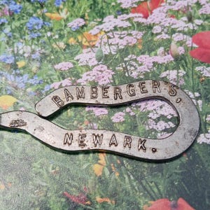 May include: A vintage, silver-toned bottle opener with the text "BAMBERGER'S NEWARK" embossed on it. The opener has a curved design and shows signs of wear, suggesting it is an antique. The background features a colorful floral arrangement.