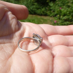 K&ouml;nnte beinhalten: Ein silberner Ring mit einem quadratischen Stein, der in einen dekorativen Rahmen gefasst ist. Der Ring befindet sich auf der Hand einer Person.