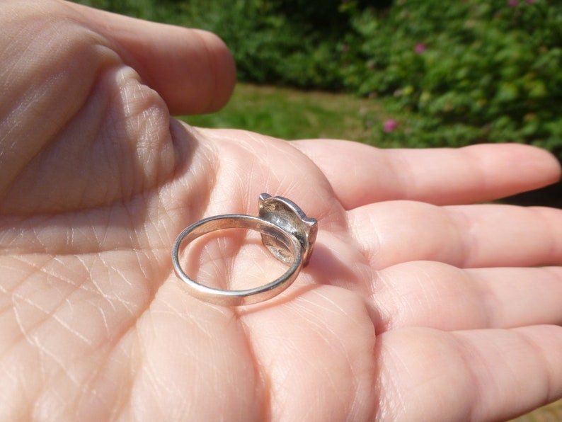 K&ouml;nnte beinhalten: Ein silberner Ring mit einem quadratischen Stein, der in einen dekorativen Rahmen gefasst ist. Der Ring befindet sich auf der Hand einer Person.