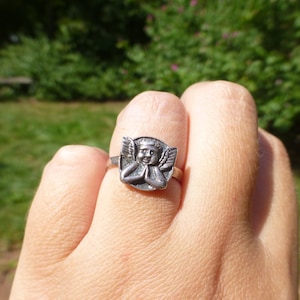 K&ouml;nnte beinhalten: Ein silberner Ring mit einem detaillierten Engeldesign. Der Engel ist mit Fl&uuml;geln dargestellt und betet mit gefalteten H&auml;nden.