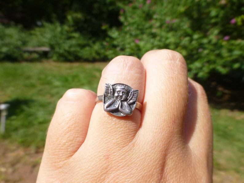 K&ouml;nnte beinhalten: Ein silberner Ring mit einem detaillierten Engeldesign. Der Engel ist mit Fl&uuml;geln dargestellt und betet mit gefalteten H&auml;nden.