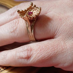 May include: A gold ring with a textured, flower-shaped design. The centre of the flower has a single, red gemstone.