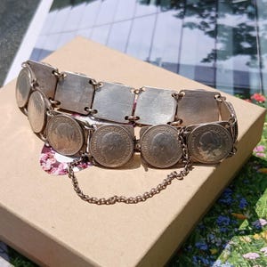 May include: A silver bracelet featuring a series of square and circular elements. The circular elements appear to be coins with a profile image. A delicate chain connects the two ends of the bracelet. The bracelet is resting on a brown cardboard box.