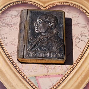 May include: A small, antique, book-shaped, metal box with a relief image of a man's face. The box is closed and has a gold-colored trim. The text "PIVS XII PONT MAX" is engraved on the front of the box.