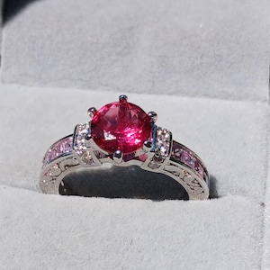 May include: A silver ring with a large, round, bright pink gemstone as the centerpiece. The ring features smaller pink stones and clear accent stones on the band. The ring is set in a gray velvet box.