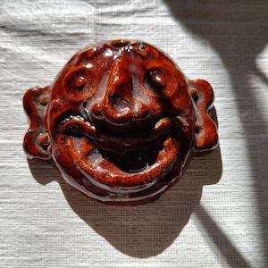 May include: A brown ceramic face mask with a wide, toothy grin. The mask has a large, bulbous nose and exaggerated features.