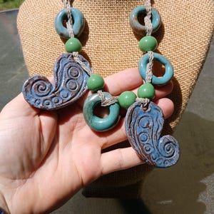 May include: A handmade necklace featuring blue ceramic heart-shaped pendants with a swirl design, teal rings, and green beads. The necklace is strung on a natural-colored cord, creating a unique and artistic accessory.