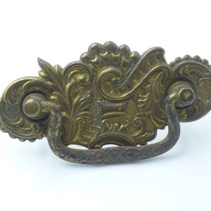 May include: Ornate brass drawer pull with a decorative floral design and a loop handle. The pull has a worn, antique finish.