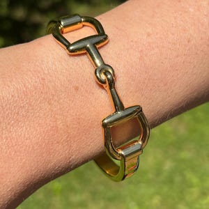 May include: A polished gold-tone metal bracelet featuring a central equestrian-inspired horse bit design. The bracelet has a smooth, reflective surface and a hinged construction that secures around the wrist with a sleek, metallic finish.