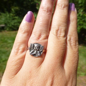 K&ouml;nnte beinhalten: Ein silberner Ring mit einem detaillierten Cherub-Design. Der Cherub tr&auml;gt eine Krone und hat die H&auml;nde zum Gebet gefaltet.