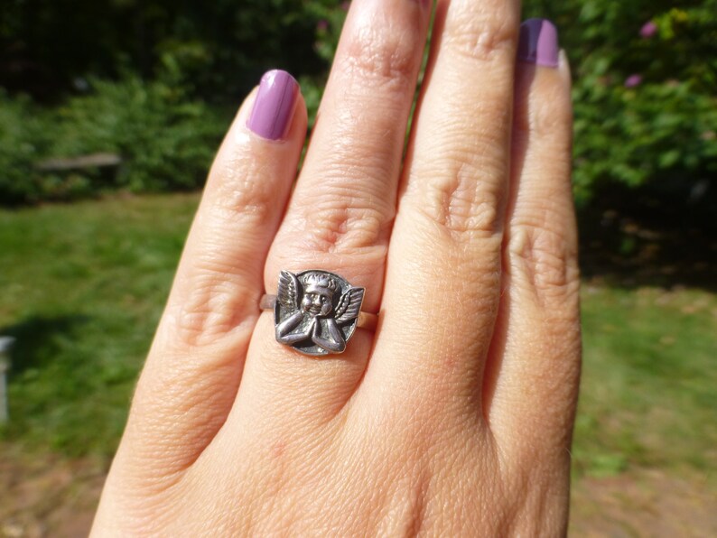 K&ouml;nnte beinhalten: Ein silberner Ring mit einem detaillierten Cherub-Design. Der Cherub tr&auml;gt eine Krone und hat die H&auml;nde zum Gebet gefaltet.