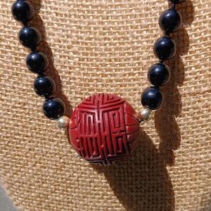 May include: A necklace featuring black, round beads and a large, carved red pendant. The pendant has an intricate geometric design. The necklace is displayed on a tan, textured surface.