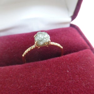 May include: A gold ring with a rough, grey diamond set in the center. The ring is in a red velvet ring box.