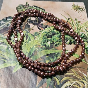 May include: A multi-strand necklace composed of dark brown, irregularly shaped pearls. The necklace is draped over a botanical print background featuring green leaves and foliage. The pearls have a lustrous sheen.