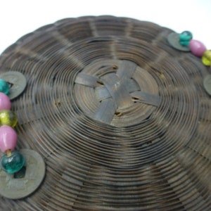 May include: A round woven basket with a brown finish. The basket has two rows of decorative beads in green, pink, and yellow. The beads are attached to the basket with metal coins.