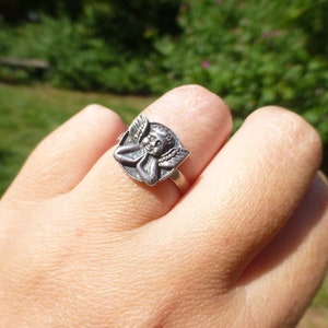 K&ouml;nnte beinhalten: Ein silberner Ring mit einem Cherub-Design. Der Cherub ist mit Fl&uuml;geln und einem l&auml;chelnden Gesicht dargestellt.