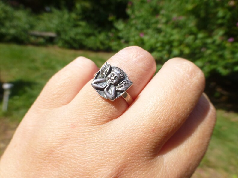 K&ouml;nnte beinhalten: Ein silberner Ring mit einem Cherub-Design. Der Cherub ist mit Fl&uuml;geln und einem l&auml;chelnden Gesicht dargestellt.