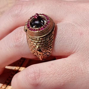 May include: A gold ring with a brick-like design and a round, red-gemstone-encrusted top. The top of the ring features a small, black, three-dimensional figure inside a clear glass dome.