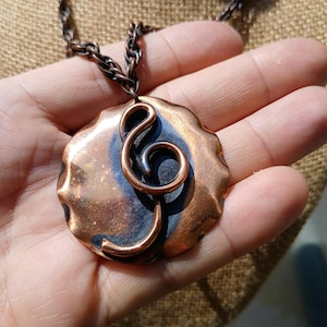 May include: A copper-colored pendant necklace featuring a stylized treble clef design. The pendant has a textured, organic shape with a dark blue-grey center. The chain is a matching dark copper color.