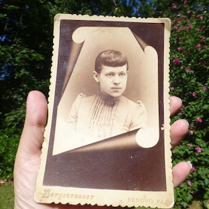 May include: A vintage black and white photograph of a young person with short hair. The photo is in a paper frame with a scalloped edge. The text "Bergotressor RENOVO, PA." is printed at the bottom of the frame.