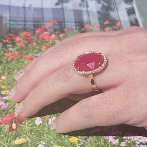 May include: A gold ring with a large, oval-shaped, red gemstone surrounded by a row of small, clear gemstones.
