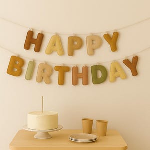 May include: A birthday celebration scene featuring a felt banner spelling "HAPPY BIRTHDAY". A cake with a candle is on a table with plates, cups, and wrapped gifts. The scene is set on a light-coloured rug.