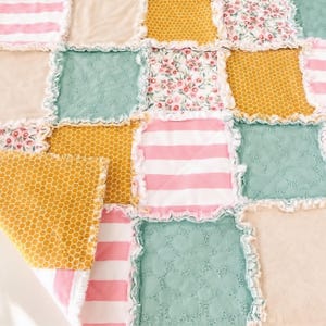 May include: A patchwork quilt featuring square fabric panels with frayed edges. The design includes alternating patterns of pink and white stripes, solid beige, mustard yellow honeycomb prints, floral motifs, and textured sage green fabric.