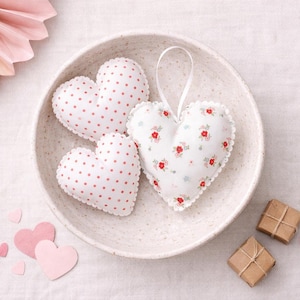 Könnte beinhalten: Drei herzförmige Dekorationen in einer weißen Schale. Zwei Herzen haben einen weißen Hintergrund mit roten Punkten, und das dritte hat ein Blumenmuster. Kleine rosa Papierherzen und verpackte Geschenke sind ebenfalls vorhanden.