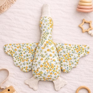 Puede incluir: Un juguete de peluche con forma de ganso y estampado floral con flores amarillas y blancas sobre un fondo blanco. Las alas están extendidas y los pies son de color beige claro. El juguete está diseñado para bebés y niños pequeños.