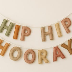 Könnte beinhalten: Ein zweireihiges Banner mit den Worten "HIP HIP HOORAY" in bunten, genähten Buchstaben. Die Buchstaben sind in Grün-, Orange-, Rosa- und Gelbtönen gehalten und an einer weißen Schnur befestigt. Der Hintergrund ist ein schlichtes Off-White.