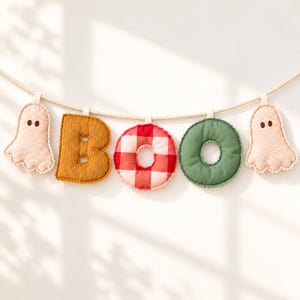 May include: A decorative banner featuring two beige ghost-shaped fabric ornaments flanking the letters B and O, which are spelled out in a mustard yellow corduroy B, a red and white checkered O, and a solid sage green O, all strung on a rope.