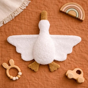 Può includere: Un peluche a forma di oca bianca con zampe e becco marroni, circondato da giocattoli di legno. Un arcobaleno di legno, un'auto e un massaggiagengive a forma di coniglietto sono disposti su una superficie color ruggine. L'oca ha le ali spiegate.