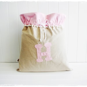 Könnte beinhalten: Großer beigefarbener Stoff-Aufbewahrungsbeutel mit rosa Oberteil und einem rosa Buchstaben "H"-Applikation. Der obere Rand ist gerafft und mit weißen Pompons verziert. Weißer Kordelzugverschluss.