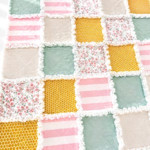 May include: A patchwork rag quilt featuring a grid of square fabric panels in alternating patterns and colours, including pink and white stripes, solid sage green, mustard yellow with a white honeycomb print, and a floral pattern with pink flowers.