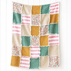 May include: A patchwork quilt featuring a grid of square fabric panels in alternating patterns and colors. The design includes mustard yellow honeycomb prints, solid beige squares, sage green panels, pink and white stripes, and floral prints.
