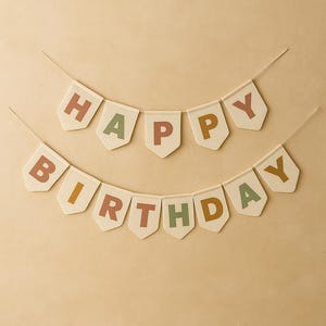 Könnte beinhalten: Zwei cremefarbene Bannergirlanden mit den Worten "HAPPY BIRTHDAY" in bunten Buchstaben. Die Buchstaben sind in Grün-, Orange- und Brauntönen gehalten. Die Banner sind an dünnen, hellfarbigen Schnüren aufgereiht, ideal für eine Geburtstagsfeier.