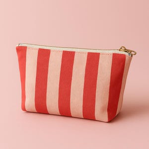 May include: A rectangular cosmetic bag with a red and light pink striped pattern. The bag has a brass-colored zipper and a light-colored interior. The bag is made of a canvas-like material and is suitable for storing makeup or toiletries.