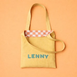 Pode incluir: Bolsa tote amarela com o nome "LENNY" em letras azuis. A bolsa tem um forro xadrez pêssego e branco. As alças e o corpo da bolsa são de cor amarela lisa. O fundo é de cor pêssego lisa.