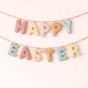 Puede incluir: Una pancarta festiva que deletrea "HAPPY EASTER" con letras de tela en colores pastel. Las letras están suspendidas de una cuerda con pequeñas pinzas para la ropa sobre un fondo blanco. Las letras son rosas, amarillas, melocotón y azules.