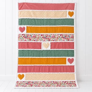 May include: A rectangular quilt featuring horizontal stripes in shades of pink, cream, green, and mustard yellow. The design includes floral patterned fabric sections and several appliqued heart shapes in solid pink, yellow, and cream colors.