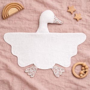 May include: A white goose-shaped baby bib with floral patterned feet and beak, set on a pink surface. Wooden toys, including a rainbow arch and star shapes, are arranged around the bib. The bib has a scalloped edge.
