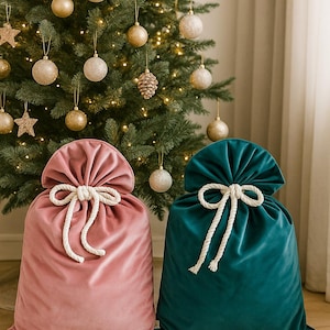 May include: Two velvet gift bags, one dusty rose and one teal, sit in front of a decorated Christmas tree. Each bag is tied with a thick, white rope. The tree is adorned with gold ornaments and lights.
