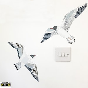 May include: Two black and white stylised illustrations of seagulls in flight against a white background. The seagulls are facing opposite directions. A white light switch is visible in the background.