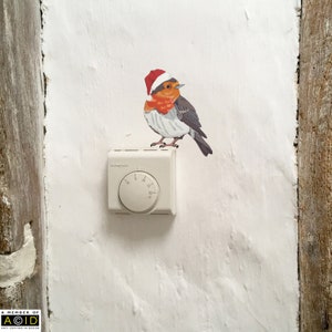 May include: A white thermostat with a round dial and a red-capped robin bird sticker on a white wall.