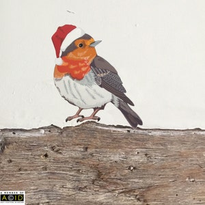 May include: A red robin wearing a Santa hat perched on a weathered wooden branch. The robin has a white chest, orange breast, and gray wings.
