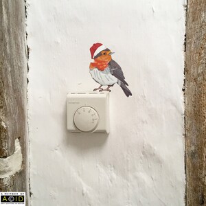 May include: A white thermostat with a round dial and a red-capped robin bird sticker on a white wall.