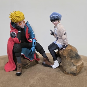 May include: Two anime-style figurines. One figure wears a red coat with blue accents, orange glasses, and yellow hair. The other figure wears a blue beret, a white jacket, and black pants. Both figures are seated on rock-like structures.