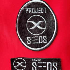 Op de afbeelding: Twee geborduurde patches in zwart en wit met de tekst "PROJECT SEEDS" en een gestileerd "X"-symbool. De grotere patch is rond en de kleinere is rechthoekig.