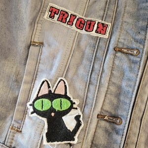 K&ouml;nnte beinhalten: Hellblaue Jeansjacke mit dem Wort "TRIGUN" in roter Schrift auf einem Aufn&auml;her. Ein schwarzer Katzenaufn&auml;her mit gr&uuml;nen Augen und wei&szlig;em Bauch ist ebenfalls sichtbar. Die Jacke hat Metallkn&ouml;pfe und ein klassisches Design.